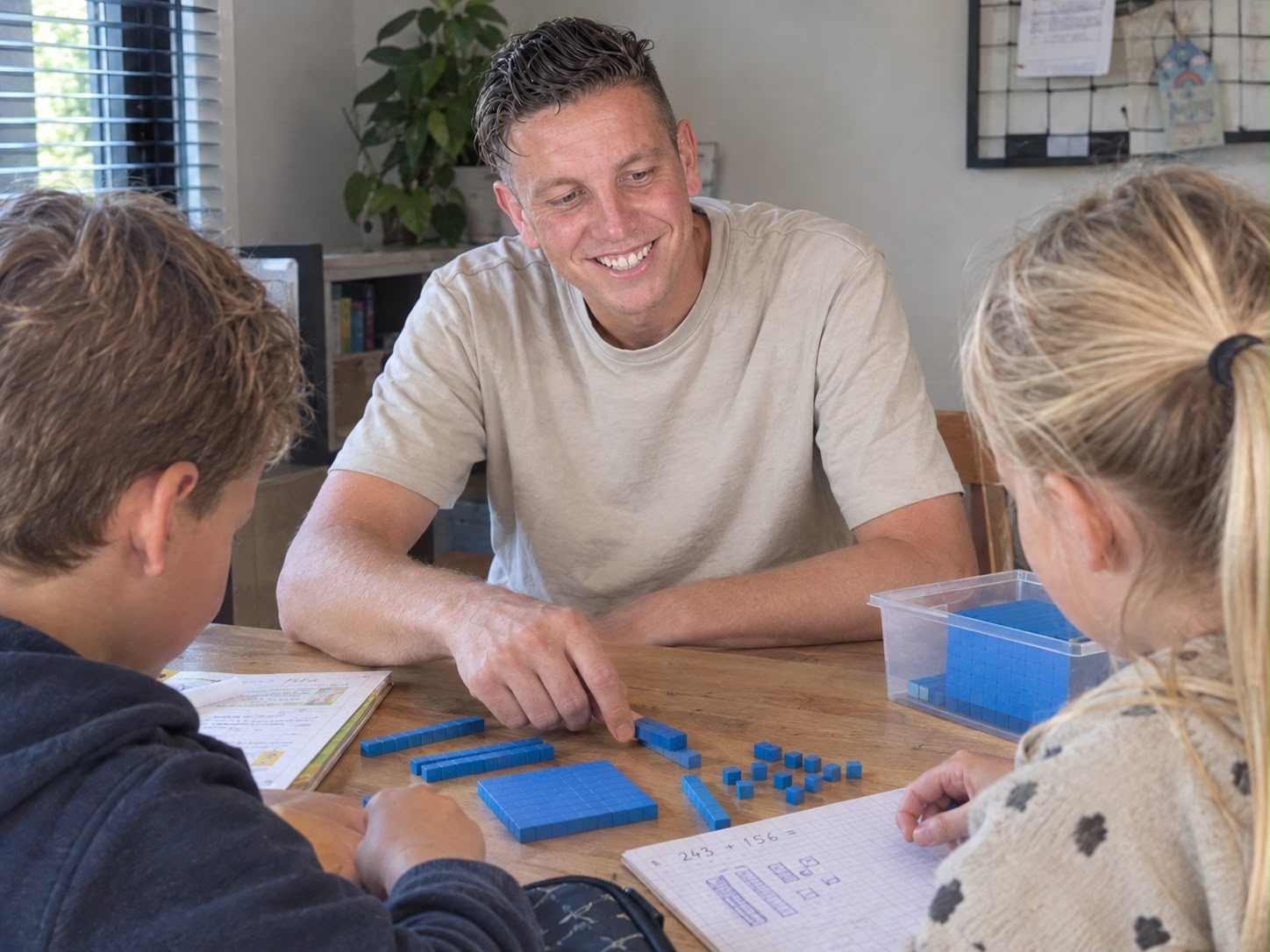 De docent legt met blauwe rekenblokjes een som uit aan twee kinderen aan de keukentafel